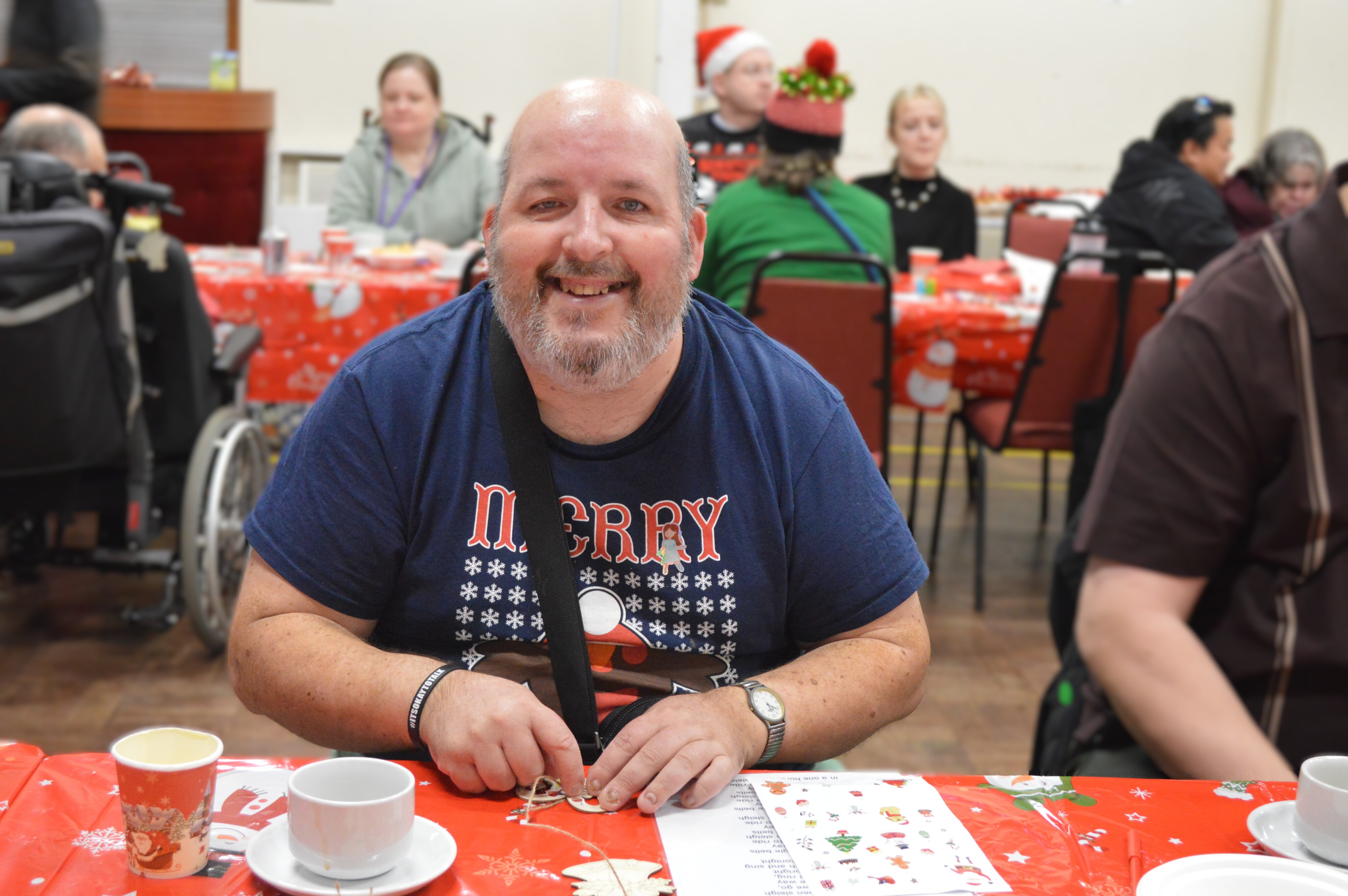 Dressed in a navy blue Christmas-themed shirt sitting at a table with red holiday tablecloth, engaging in a craft activity indoors.