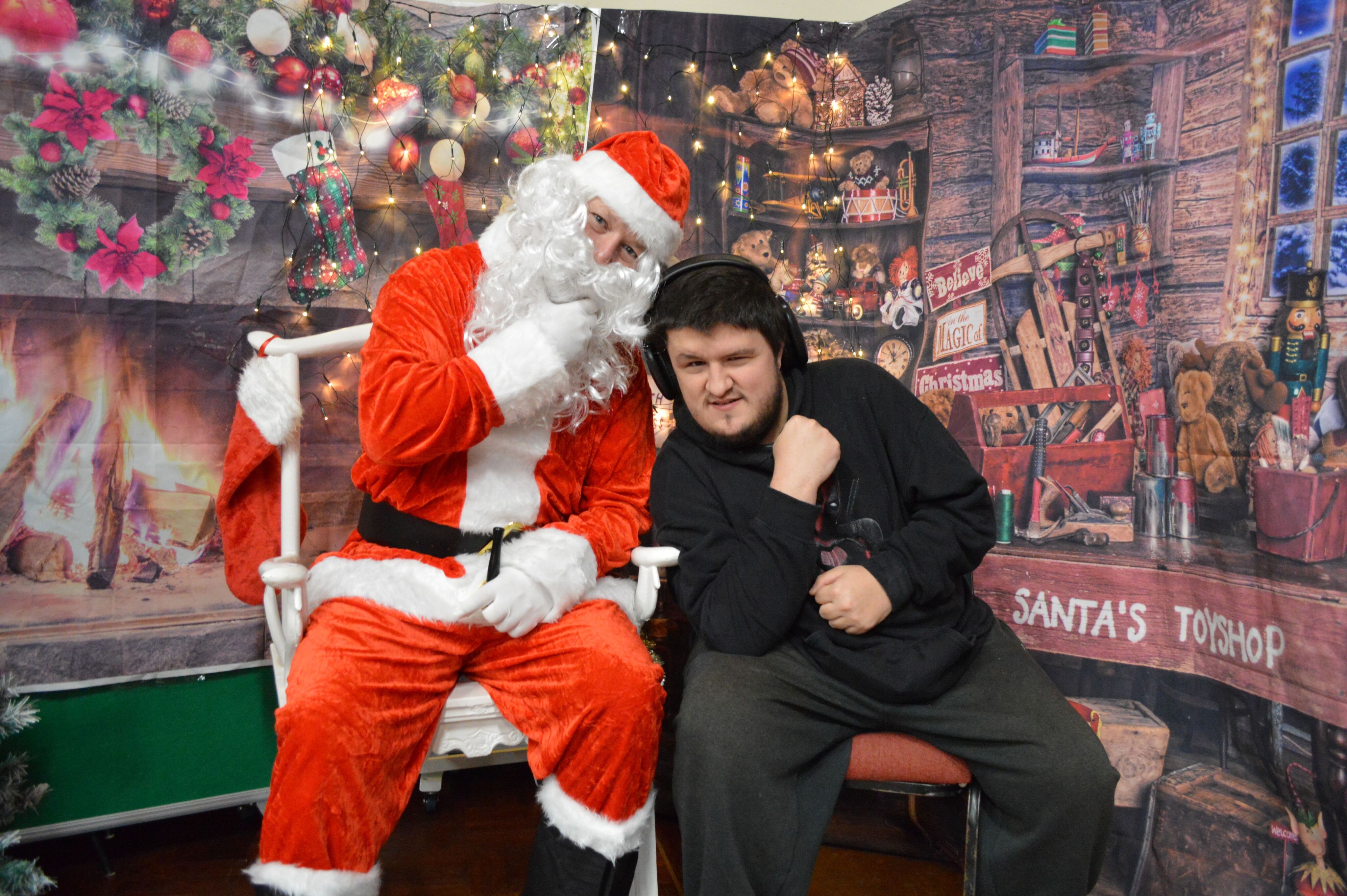 Father Christmas sitting next to a Sam in a cosy, decorated grotto with Christmas lights.