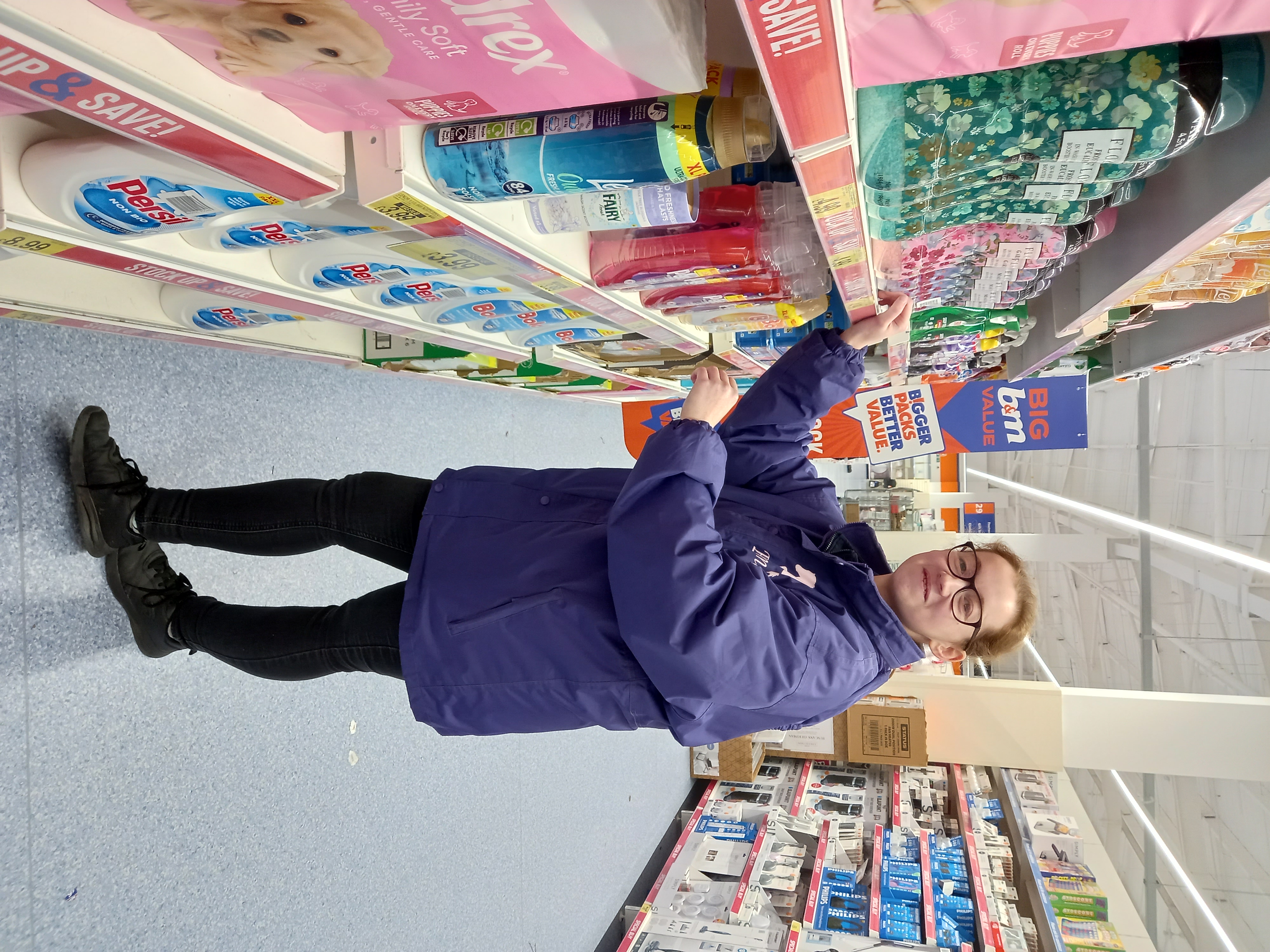 Emma selecting a product from a supermarket aisle filled with various household items.