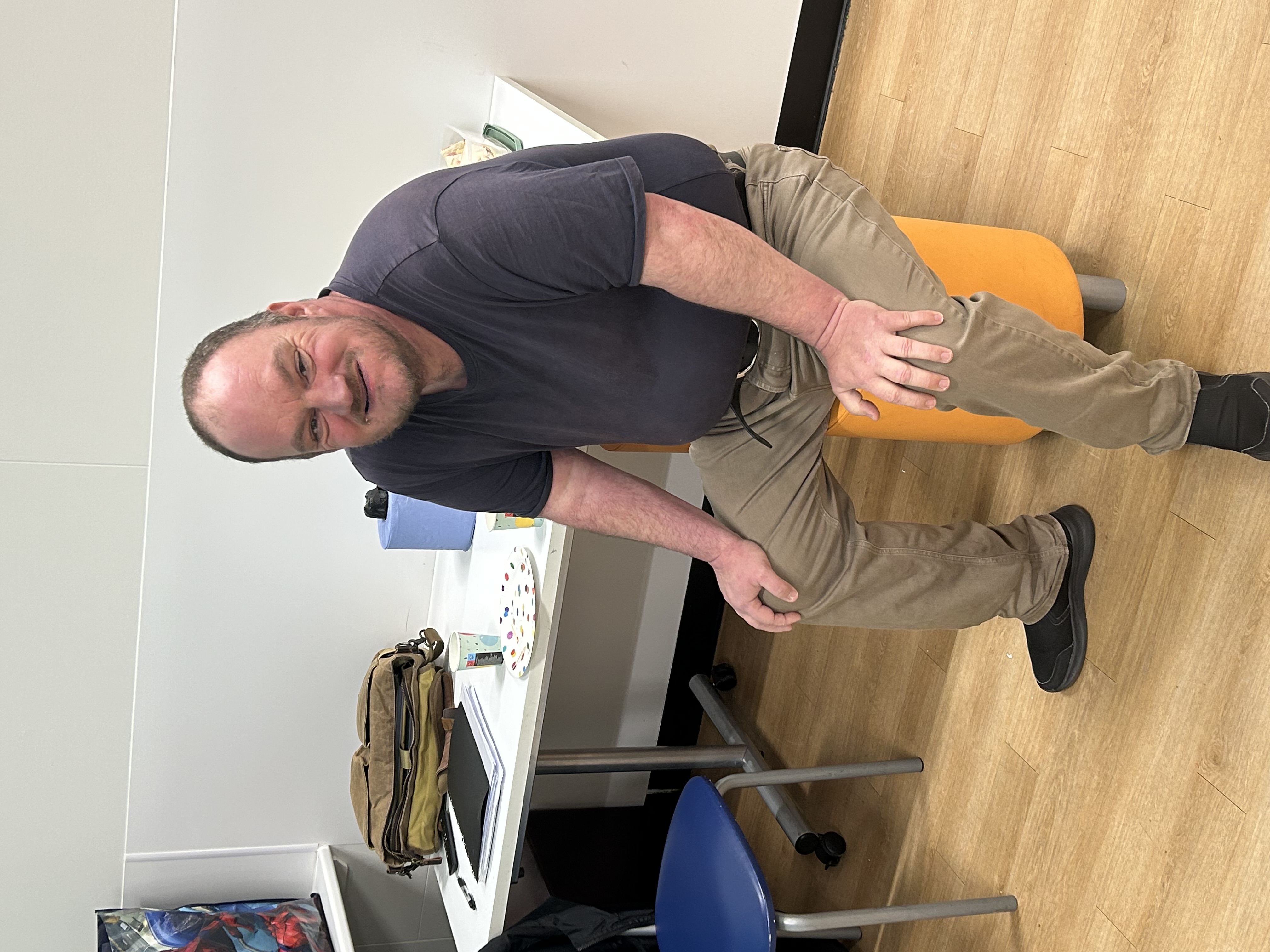 A man in a grey top and beige trousers, sitting on a yellow stool, smiling